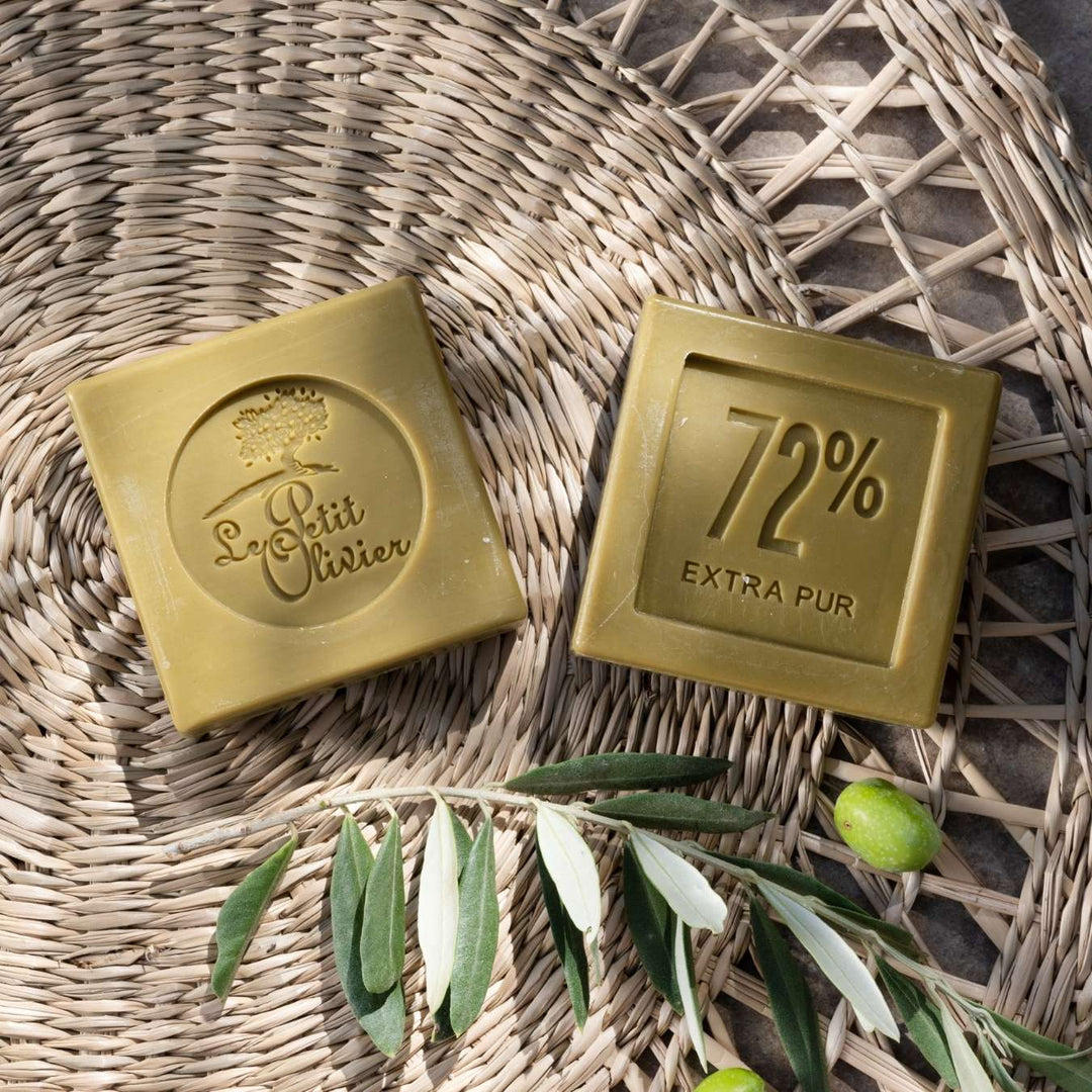 Deux pains de savon verts reposent sur un support tressé. L'un porte l'inscription « Le Petit Olivier » et le dessin d'un arbre en relief, tandis que l'autre porte la mention « 72 % EXTRA PUR ». Des feuilles d'olivier et des olives sont disposées à côté des savons.